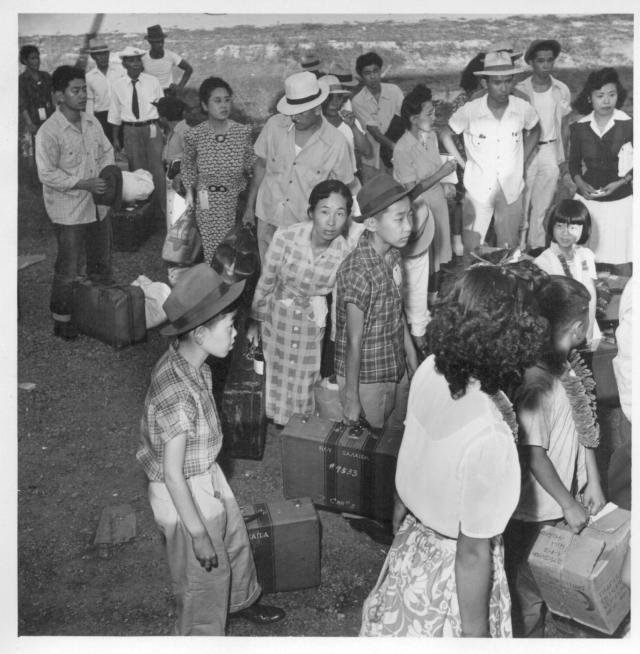 Closing of the Jerome Center, Denson, Arkansas. Jerome residents with their hand luggage wait at the [illegible] car entrance for their names to be called by the W.R.A. official checking the list. -- Photographer: Iwasaki, Hikaru -- Denson, Arkansas. 6/18/44. Source.