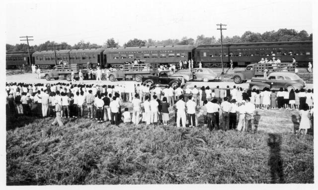 Four trucks (three can be seen in this picture) were used to transport segregees from their blocks to each car on the first segregation train (trip 4) to leave this center for Tule Lake. Large crowd of center residents (foreground) came to watch the train off but stayed inside the boundary fence on this side of the road. With the segregees came their hand luggage. -- Photographer: Lynn, Charles R. -- Dermott, Arkansas. 9/15/43. Source.