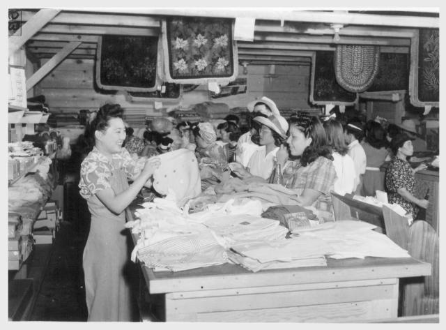 Tule Lake, Newell, Calif.--Lillian Hiraike, 18, evacuee from Seattle, Washington, shows a sweater to a prospective buyer in the general store at this War Relocation Authority center for evacuees of Japanese ancestry. -- Photographer: Stewart, Francis -- Newell, California. 7-1-42. Source.
